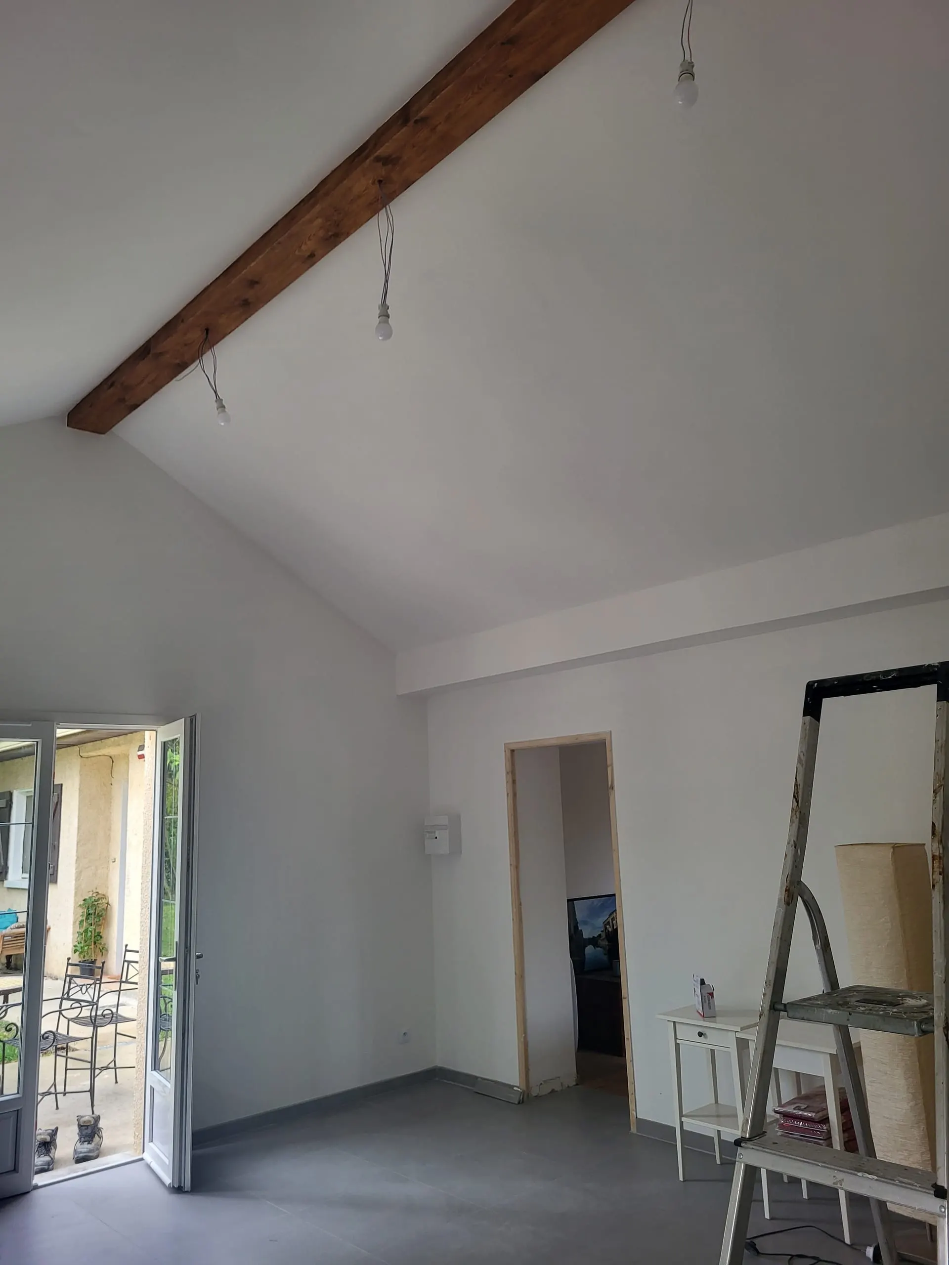 Pièce en cours de rénovation avec poutre en bois apparente, murs blancs et carrelage gris.