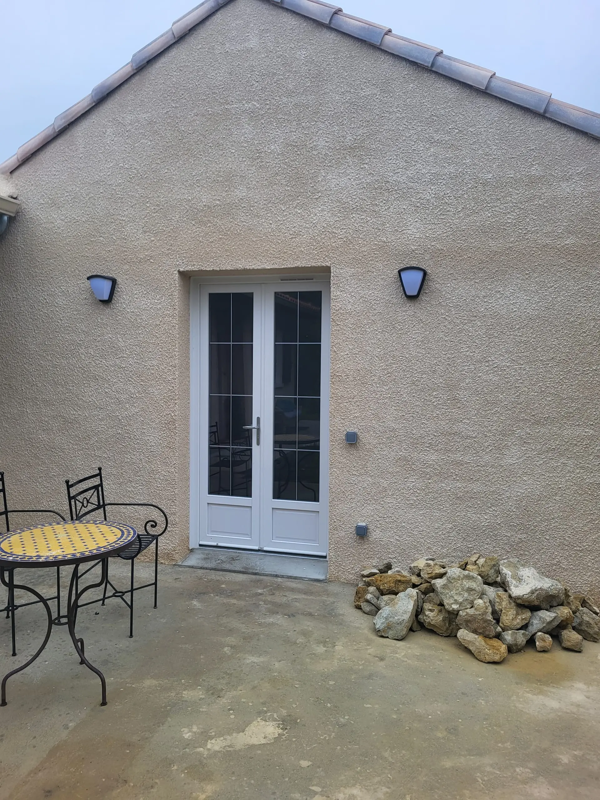 Façade enduite avec porte-fenêtre blanche et pierres posées sur une dalle en béton.