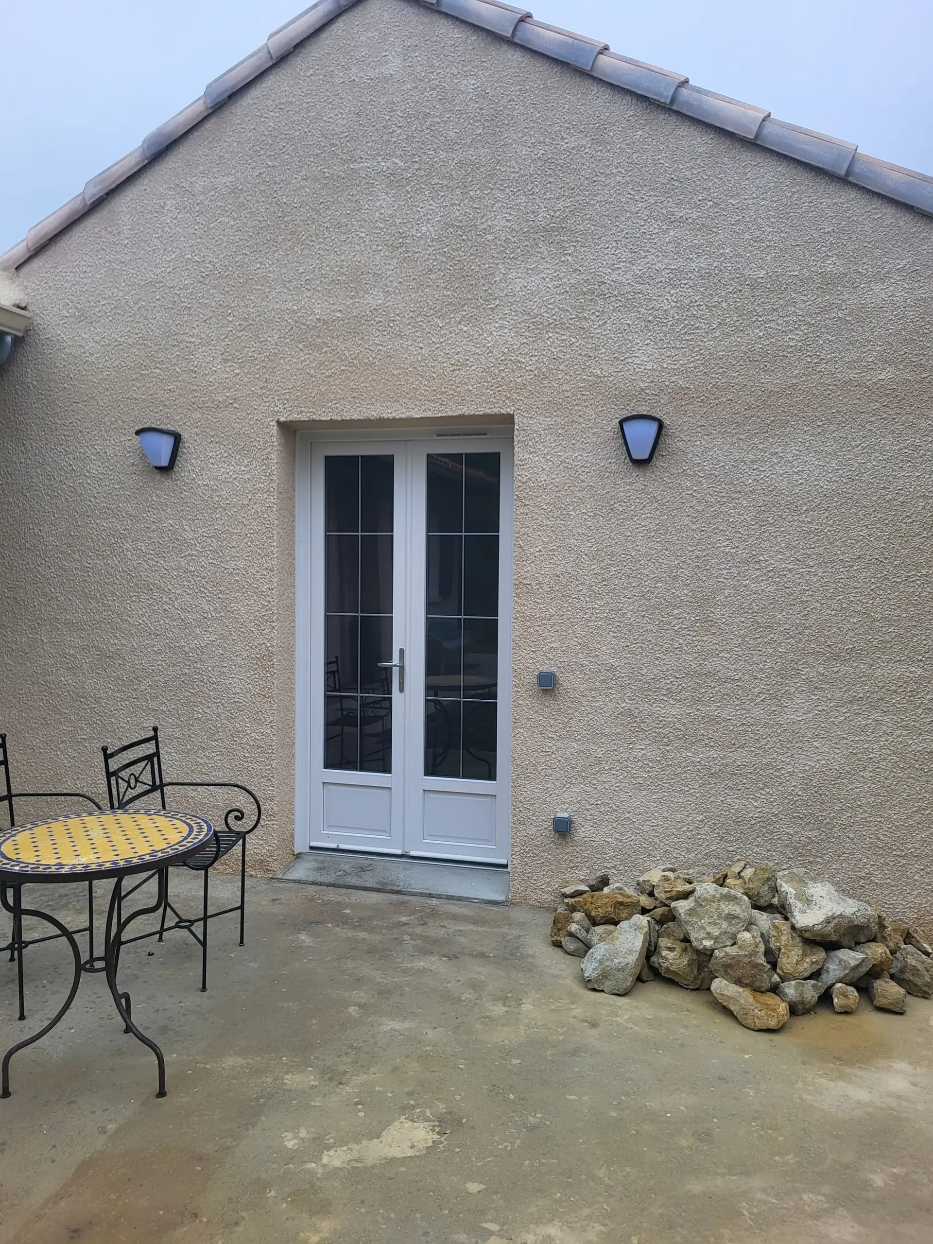 Façade enduite avec porte-fenêtre blanche et pierres posées sur une dalle en béton.